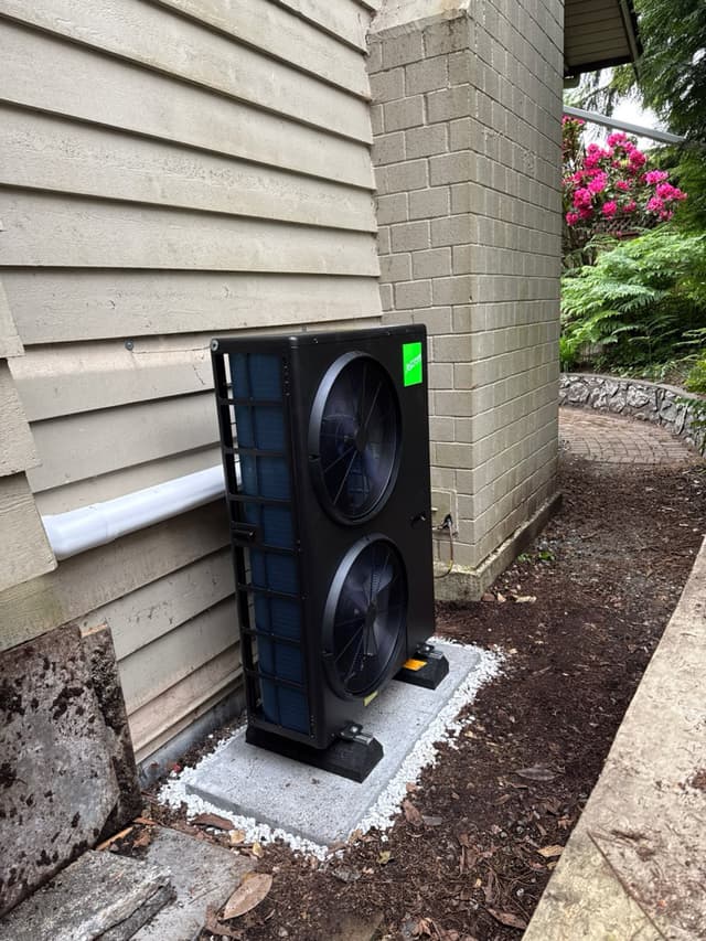 Black Jetson Air 4-ton heat pump on a concrete pad with white gravel, set between lap siding and a brick column in a North Vancouver backyard garden with blooming pink rhododendrons in the background.