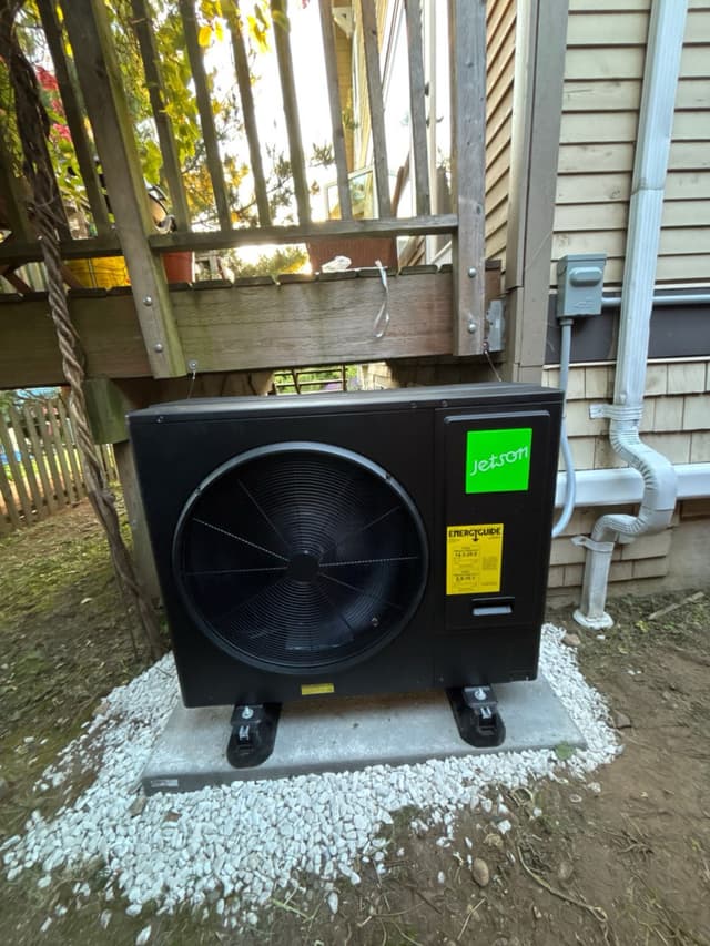 Black Jetson Air heat pump condenser on a concrete pad with white gravel, installed beneath a wooden deck against the side of a North Vancouver home, with line-set conduit running up to an electrical disconnect box.