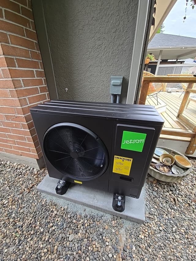 Jetson Air 3-ton heat pump mounted on a concrete pad in a river-rock gravel bed beside a brick-and-stucco house in Edgemont Village, North Vancouver, with a wooden deck and stone planter visible to the right.