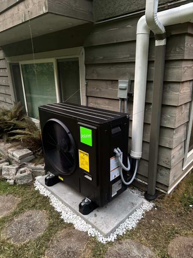 Black Jetson Air 3-ton central ducted heat pump installed against weathered wood siding at a Lynn Valley, North Vancouver home, with refrigerant lines running up the wall to a disconnect box.