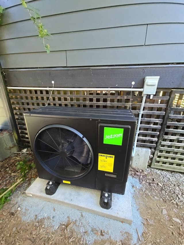 A black Jetson Air 3-ton heat pump on a concrete pad against the grey-sided exterior of a home in Lynn Valley, North Vancouver, with an electrical disconnect box mounted to the right on a lattice fence panel.