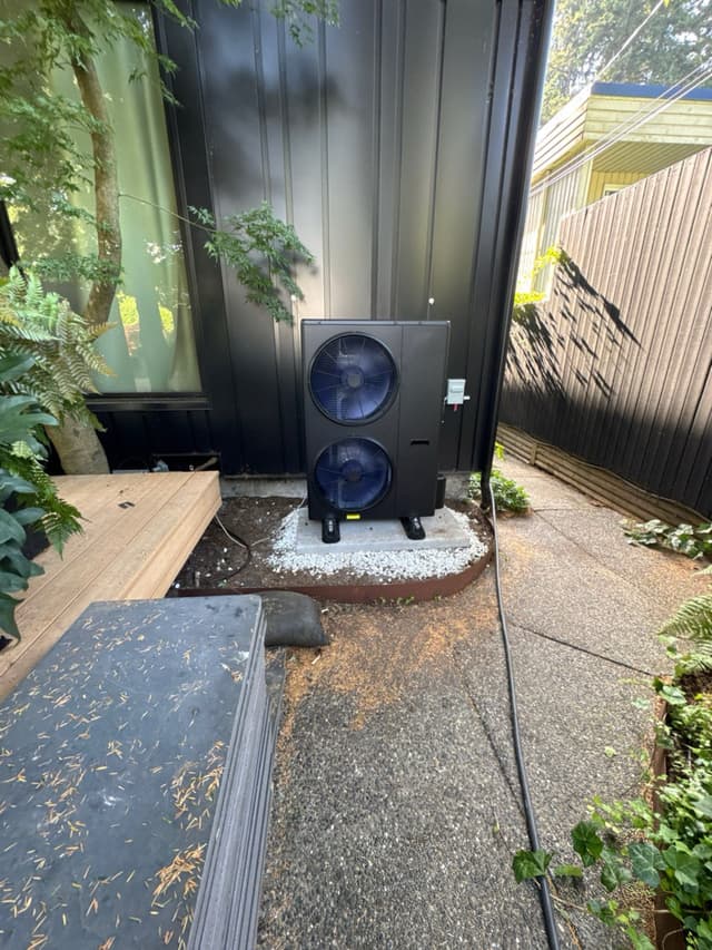 Black dual-fan 4-ton heat pump condenser installed against the black metal exterior wall of a modern home in Lions Gate, Lower Capilano, North Vancouver, with white gravel ground cover, a wooden deck, and a cedar fence visible nearby.