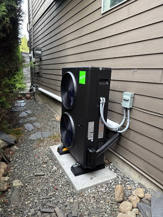 Black Jetson Air 4-ton heat pump condenser installed along the side of a taupe house in Edgemont Village, North Vancouver, with evergreen shrubs and a gravel pathway alongside.