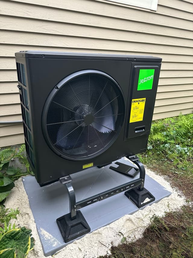 Black Jetson Air 3-ton central ducted heat pump outdoor unit installed against beige vinyl siding with green ferns on either side, Northborough, MA.