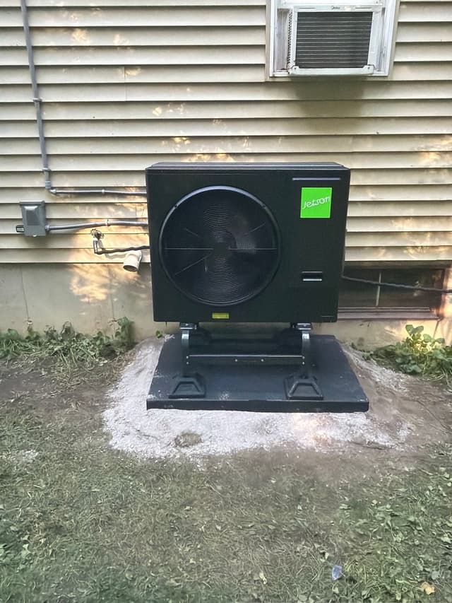 Installed black Jetson Air 3-ton central ducted heat pump condenser against tan vinyl siding of a home in Northbridge, MA.
