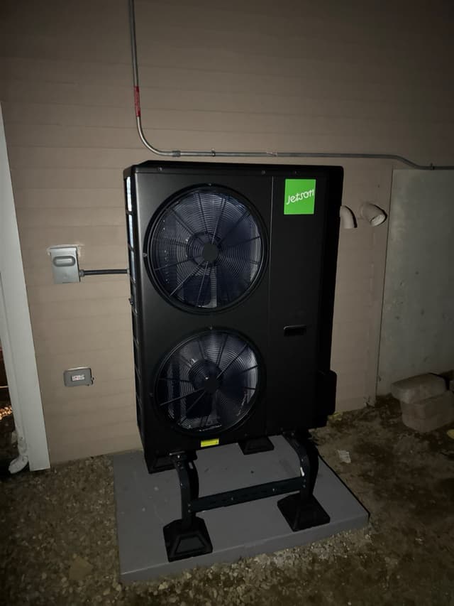 A black Jetson Air dual-fan heat pump condenser on a metal riser stand on a concrete pad beside a beige vinyl-sided house in Norton, MA, with a disconnect box and conduit visible on the wall.