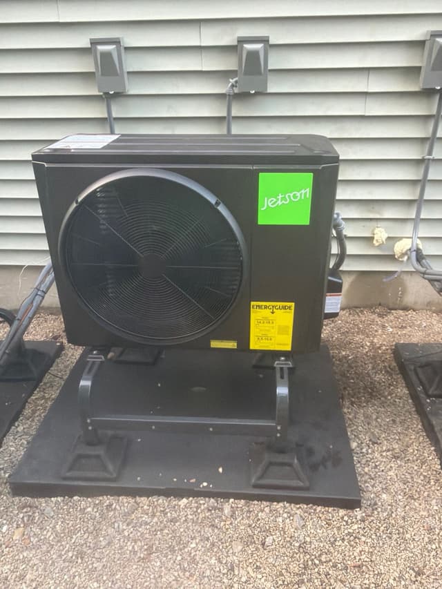 Jetson Air 2-ton heat pump condenser mounted on a black riser stand over gravel beside a light gray vinyl-sided house in Norton, MA, with line-set conduit and two electrical disconnect boxes visible on the wall behind it.
