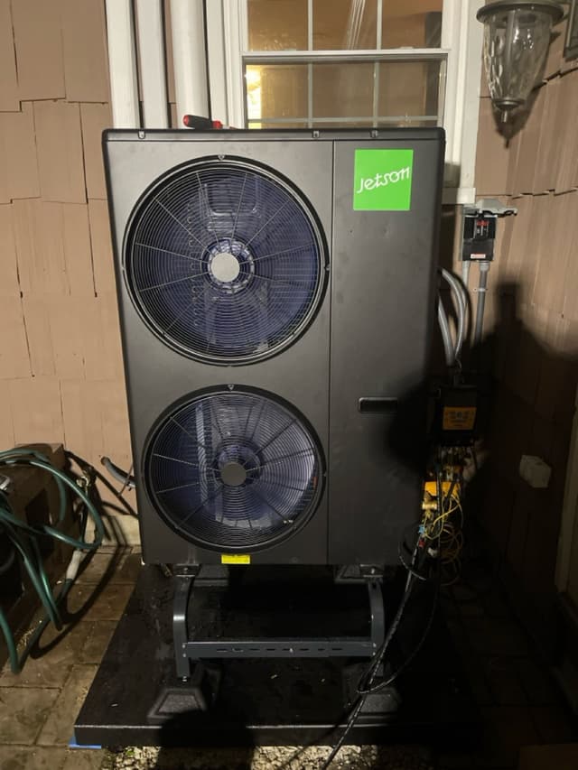 Black dual-fan 4-ton heat pump condenser on a metal stand mounted against a wood-sided house in Ossining, NY, photographed at night with electrical conduit and disconnect box visible to the right.
