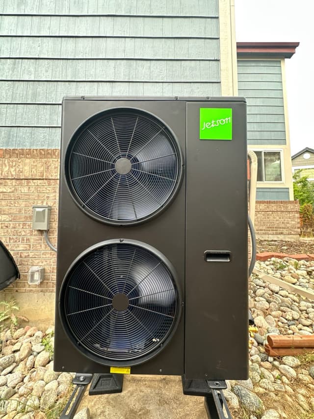 A large black Jetson Air 4-ton heat pump installed in a river-rock landscaped area along the brick and grey-blue siding exterior of a two-story home in Parker, Colorado.