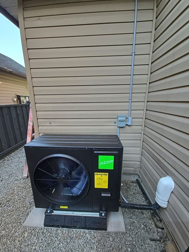 Jetson Air 3-ton central ducted heat pump unit installed in the corner between two sections of beige vinyl siding at a home in Pitt Meadows, BC, with a gray conduit running up the wall to a disconnect box and a white line-set cover to the right.
