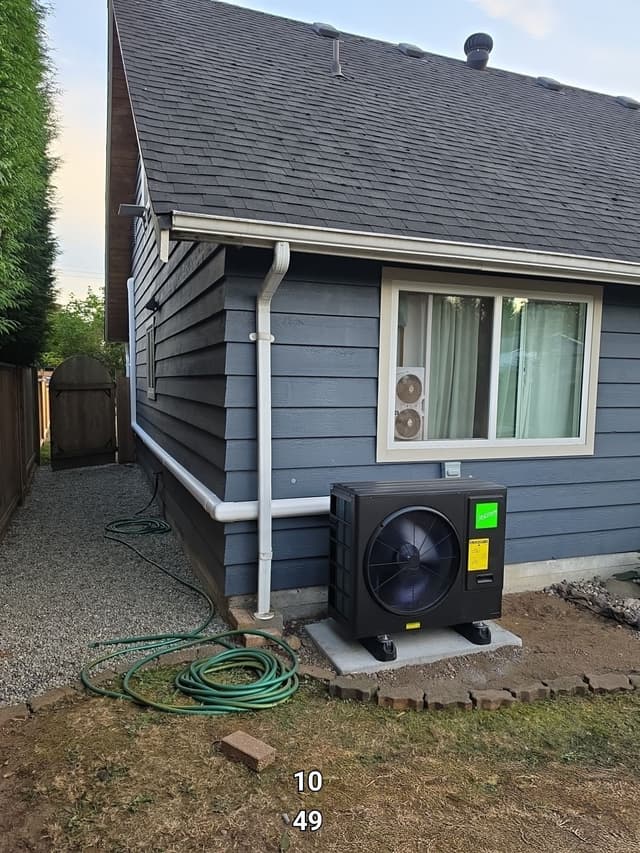 Black Jetson Air 3-ton heat pump installed along the side of a blue-grey lapped-siding house in Pitt Meadows, BC, with a white downspout on the wall and gravel and grass in the foreground.