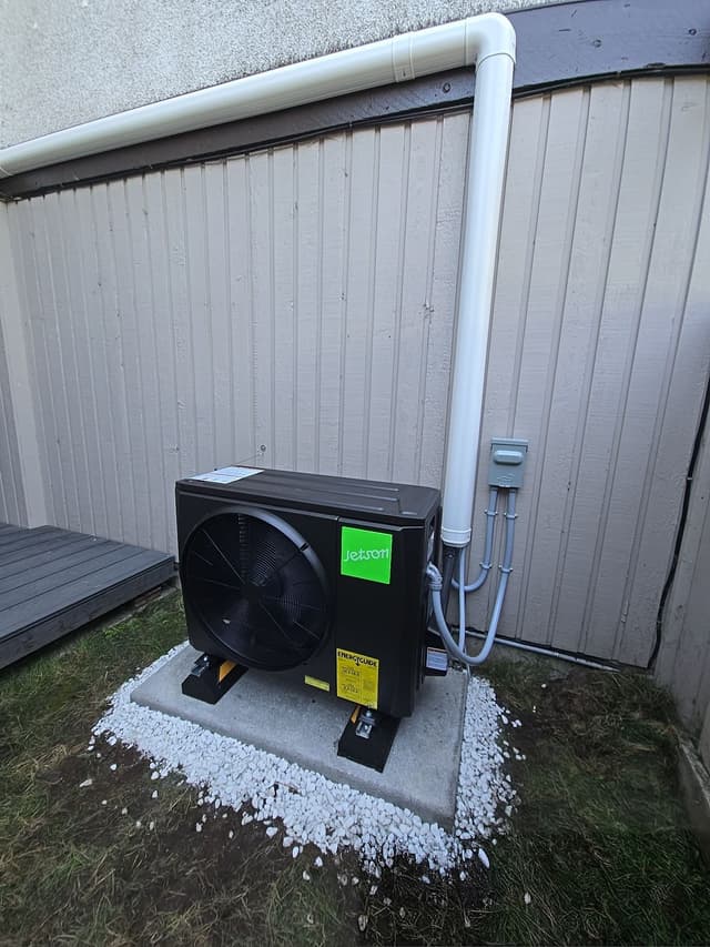 Black Jetson Air 2-ton central ducted heat pump mounted against vertical board siding with white decorative gravel, Birchland Manor, Port Coquitlam.