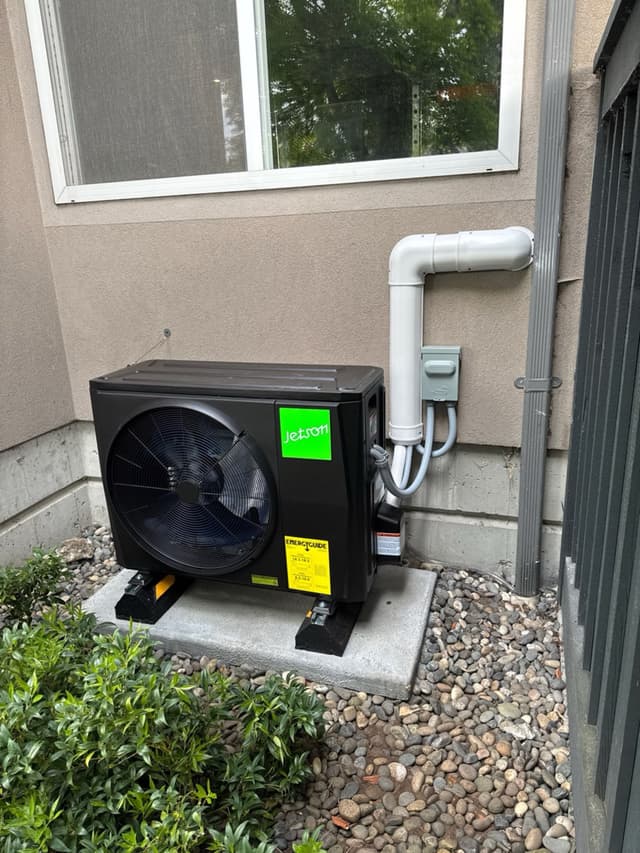 Jetson Air 2-ton heat pump condenser on a concrete pad in a gravel side yard against a beige stucco wall in Port Coquitlam, BC, with white PVC conduit running up to a grey electrical disconnect box.