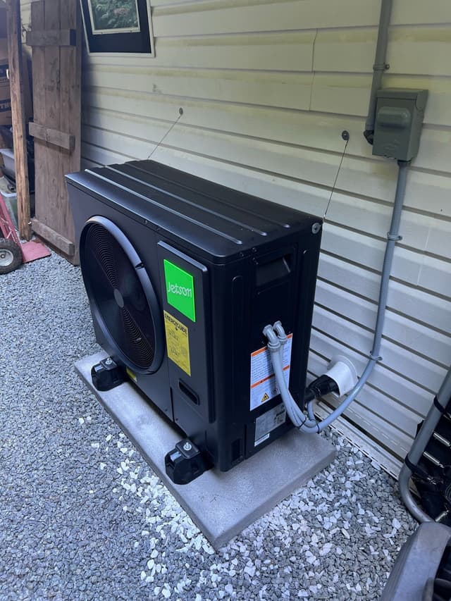 Black Jetson Air heat pump installed on a concrete pad in a gravel area beside a light-colored horizontal-siding building in Eagle Ridge, Port Moody, with refrigerant lines running into the wall and a disconnect box mounted on the siding above.