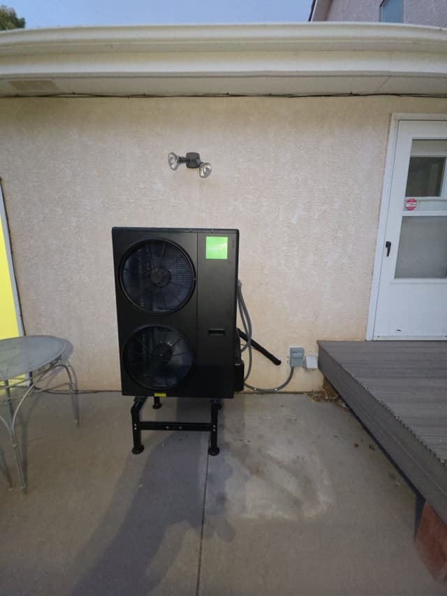 A black Jetson Air heat pump on a metal stand on a concrete patio against a stucco wall, with conduit and wiring running neatly to a wall-mounted electrical box, photographed at dusk in Pueblo, CO.