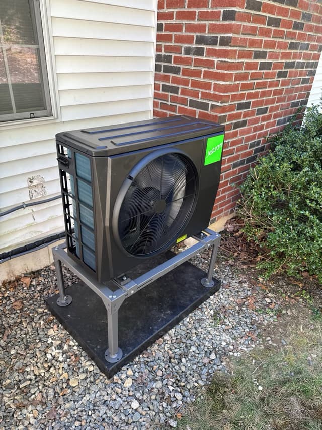 Black Jetson Air heat pump condenser installed along the side of a house with white vinyl siding and red brick in Raynham, MA.