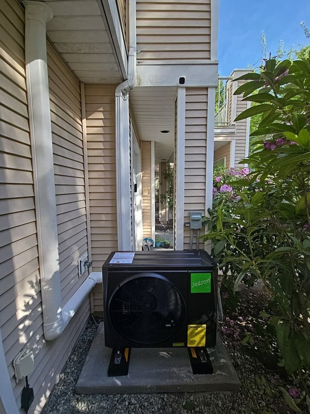 Black Jetson Air heat pump condenser on a concrete pad along the side of a beige vinyl-sided house in Richmond, BC, with white refrigerant line covers running up the wall and a pink rhododendron bush in bloom to the right.
