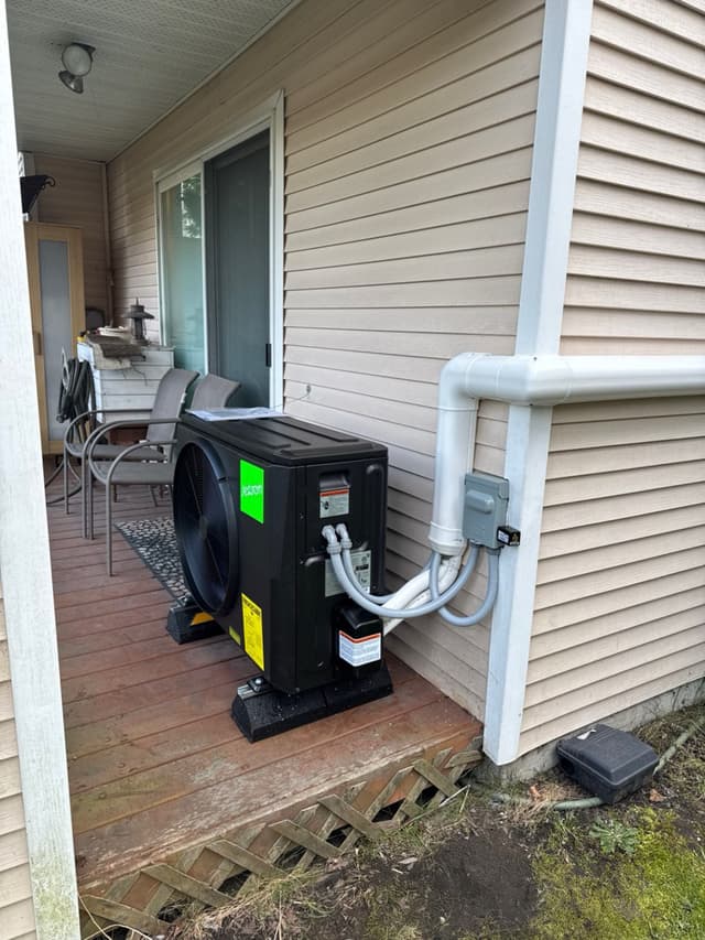 Jetson Air 2-ton central ducted heat pump mounted on rubber isolation pads on a wood deck beside a house with beige vinyl siding in Richmond, BC, with white PVC conduit running to a grey disconnect box.
