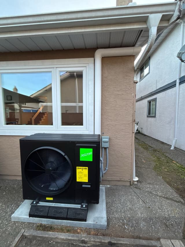 Black Jetson Air 3-ton heat pump condenser on a concrete pad with anti-vibration feet, mounted along the stucco side wall of a Richmond, BC home with conduit running neatly to a disconnect box above.