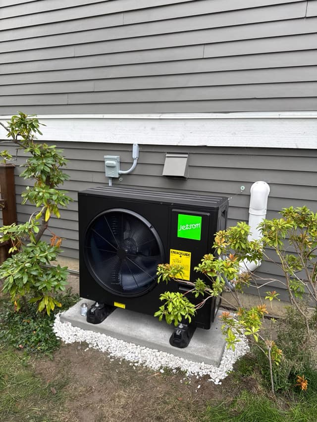 Black Jetson Air 3-ton heat pump condenser on a new concrete pad with white decorative gravel, mounted against gray lap siding with electrical disconnect and refrigerant lines run up the wall, shrubs on either side, in Steveston, Richmond, BC.