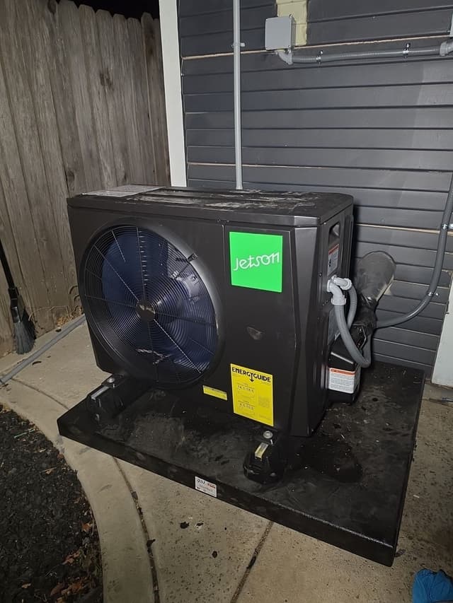 Jetson Air 2-ton heat pump condenser installed beside a gray-sided house in Land Park, Sacramento, photographed at night with the unit fully lit and conduit running up the wall behind it.