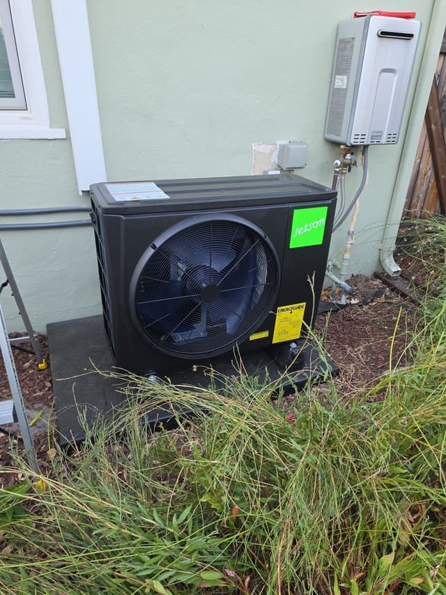 2-ton Jetson Air central ducted heat pump condenser installed on a gravel strip alongside a light green stucco house in East Sacramento, with a tankless water heater and electrical disconnect mounted on the wall to the right.