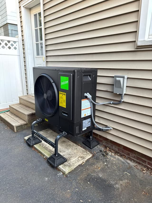 Black Jetson Air 3-ton central ducted heat pump condenser installed beside a tan vinyl-sided home in Ball Square, Somerville, with conduit running to a wall-mounted disconnect box.