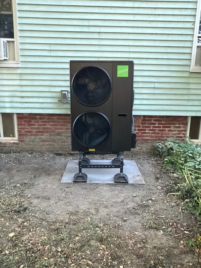 Black Jetson Air 4-ton heat pump unit installed against the light-blue lap siding and exposed brick foundation of a home in Ball Square, Somerville.