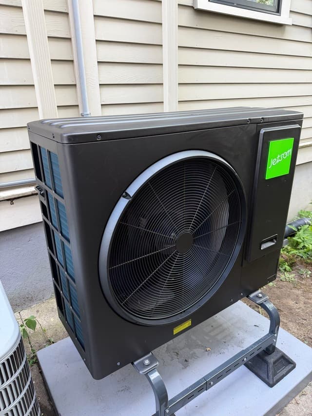 Black Jetson Air 3-ton central ducted heat pump mounted against the side of a beige house in Ball Square, Somerville, with refrigerant lines running up the wall and a second unit partially visible at the edge of the frame.