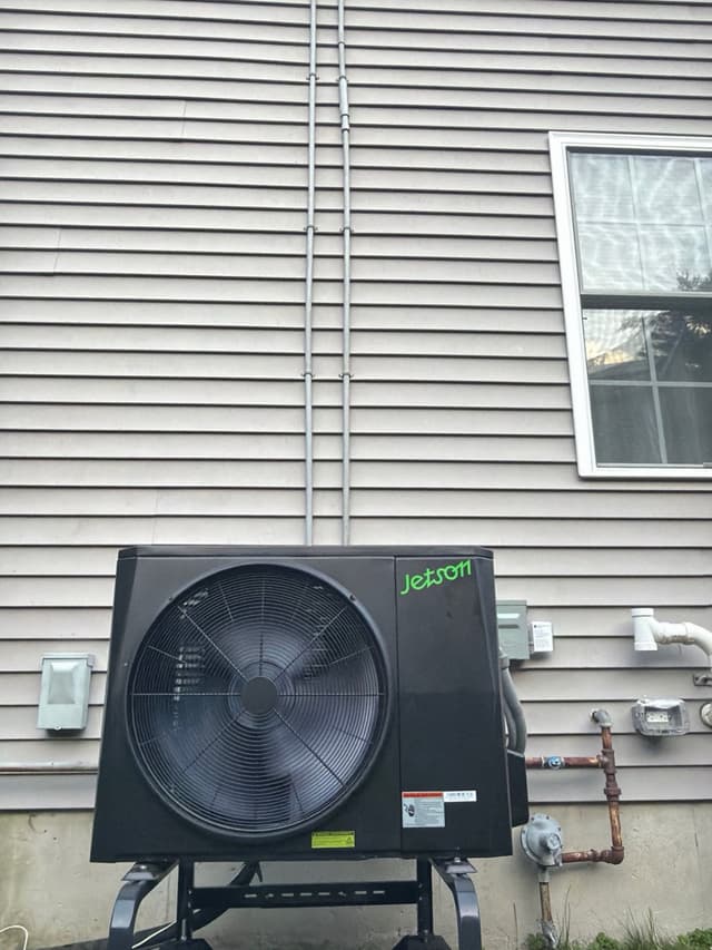 A black Jetson Air 2-ton heat pump condenser mounted on a metal stand against gray vinyl siding in Stoughton, MA, with conduit lines running up the wall and a disconnect box and gas meter visible nearby.