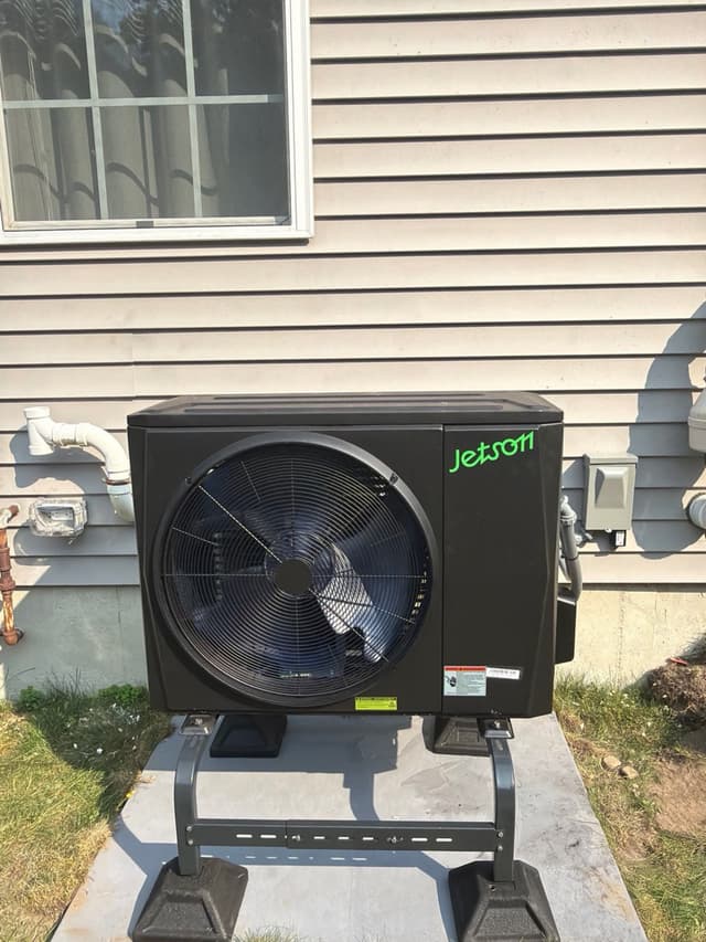 A black Jetson Air 2-ton heat pump condenser mounted on a metal stand on a concrete pad beside a house with gray vinyl siding in Stoughton, MA.