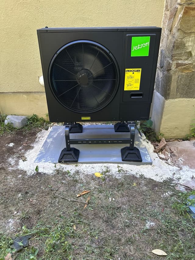 Black Jetson Air 3-ton central ducted heat pump installed against a yellow stucco wall with a stone corner in Stoughton, MA.