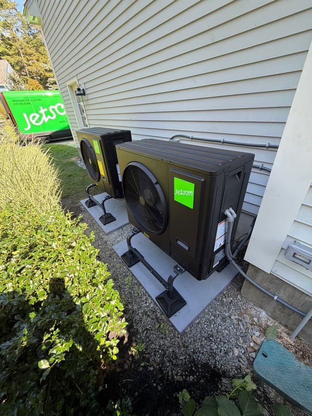Two black Jetson Air heat pump condensers installed side by side along the vinyl siding of a home in Stow, MA, with refrigerant lines running neatly along the wall.