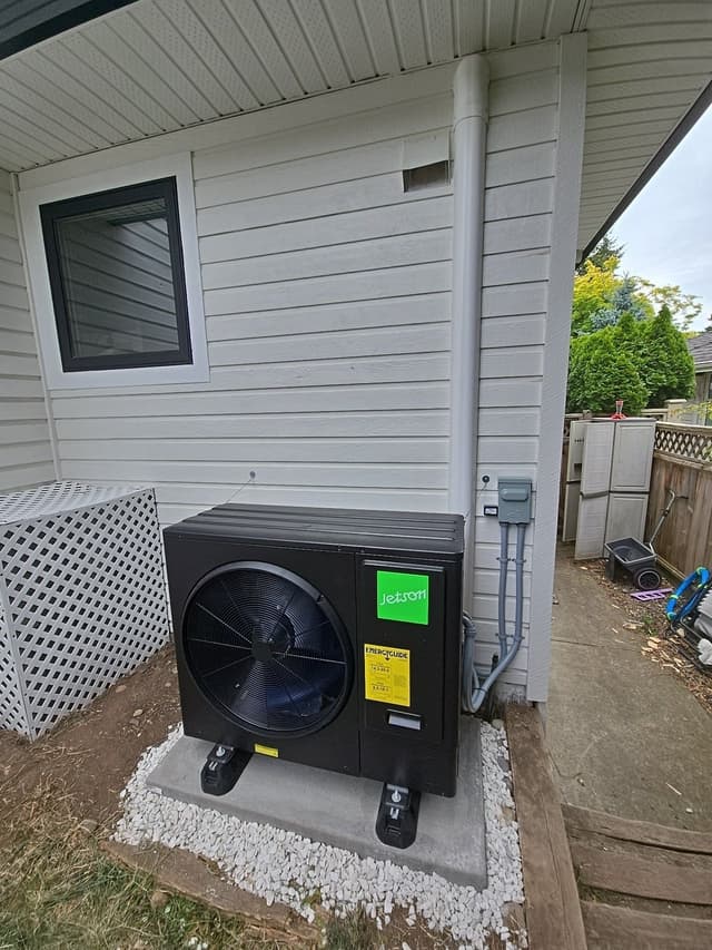 Black Jetson Air 3-ton central ducted heat pump condenser installed beside a light grey house in Ocean Park, Surrey, with conduit running up to a wall-mounted disconnect box.