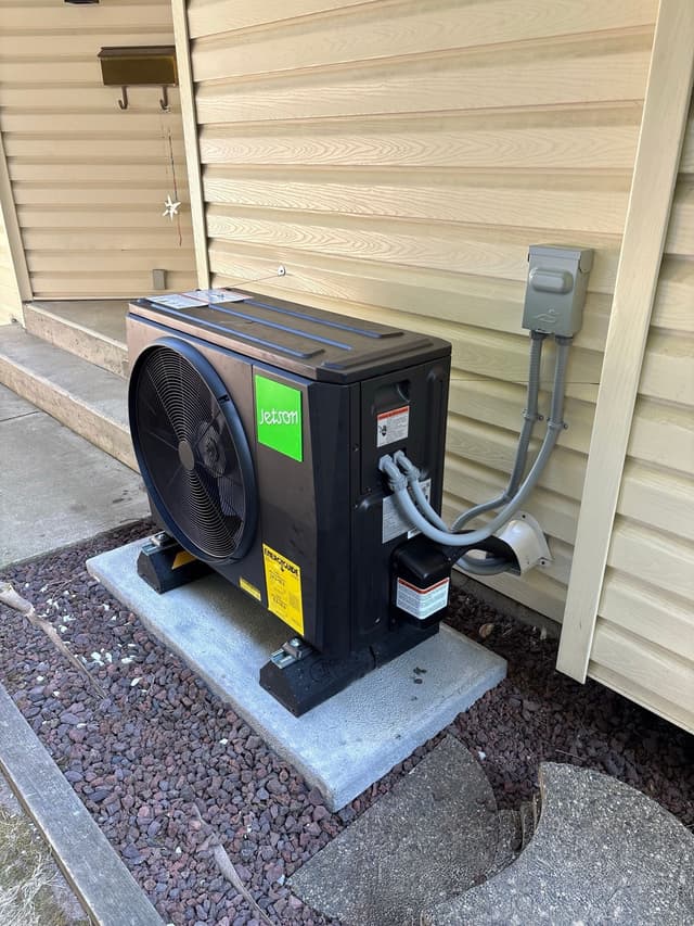 Jetson Air 2-ton heat pump condenser installed along the side of a house in Clayton Heights, Surrey, with conduit and refrigerant lines running up the wood-grain siding to a wall-mounted disconnect box.