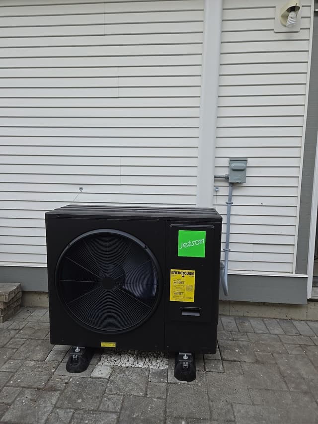 A black Jetson Air 3-ton heat pump condenser mounted on anti-vibration feet on an interlocking paver pad against the white horizontal siding of a home in Clayton Heights, Surrey, BC, with electrical conduit running up to a disconnect box on the wall.