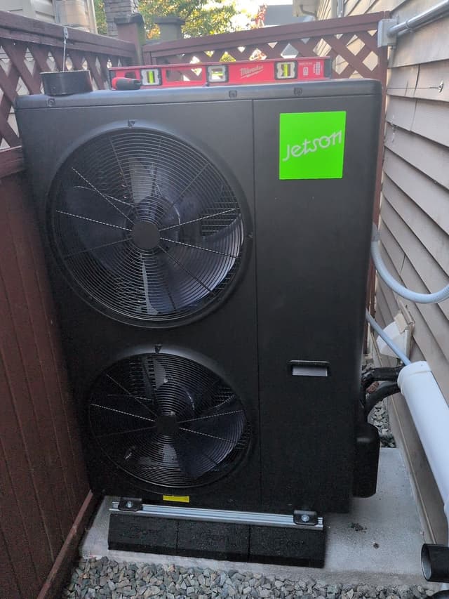 Black Jetson Air dual-fan outdoor heat pump condenser installed in a narrow side yard in Surrey Centre, Surrey, with white conduit running along the house wall and a red Milwaukee level resting on top of the unit.