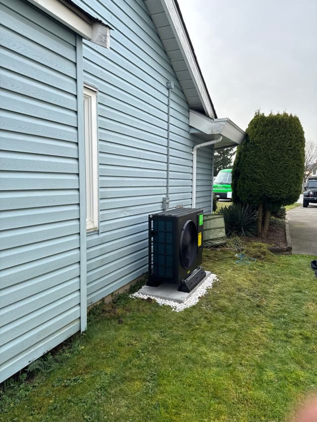 A black Jetson Air 3-ton heat pump condenser mounted on a concrete pad with white decorative gravel against a light-blue vinyl-sided house in Whalley, Surrey, BC.
