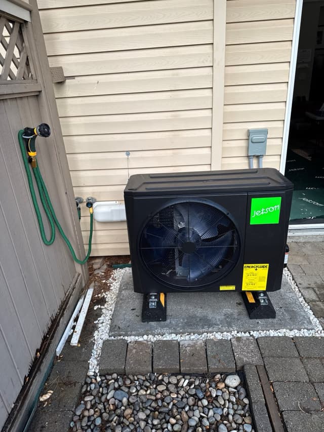 Jetson Air 2-ton heat pump mounted on a concrete paver pad with river rock surround, beside horizontal lap siding and a wooden fence in Clayton Heights, Surrey, BC.