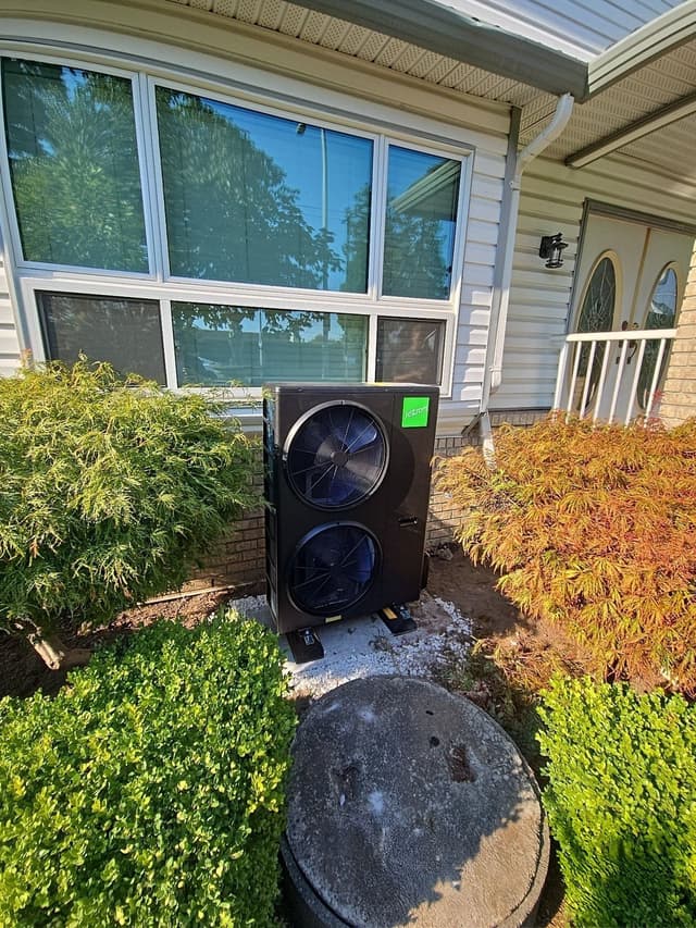 A black Jetson Air dual-fan outdoor heat pump condenser installed in a front garden bed beside ornamental shrubs, against a white vinyl-sided home in Johnston Heights, Surrey.