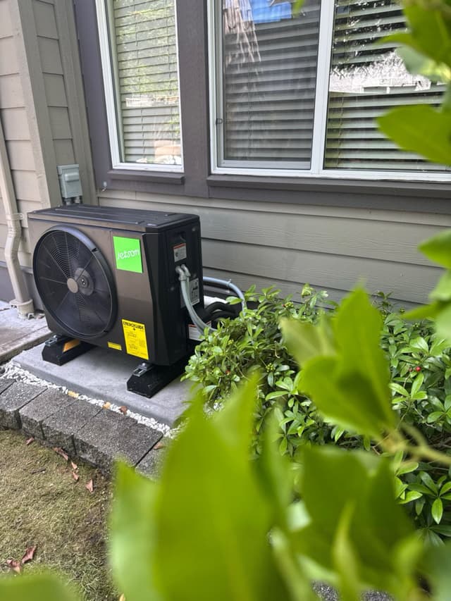 Black Jetson Air 2-ton outdoor heat pump unit on a concrete pad with white gravel and green shrubs, mounted against grey siding of a home in Sullivan, Surrey, BC.