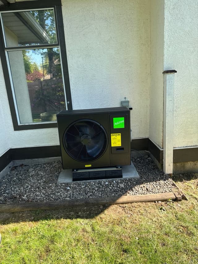 Black Jetson Air 3-ton heat pump condenser mounted on a concrete pad against a white stucco exterior wall in Fraser Heights, Surrey, BC.