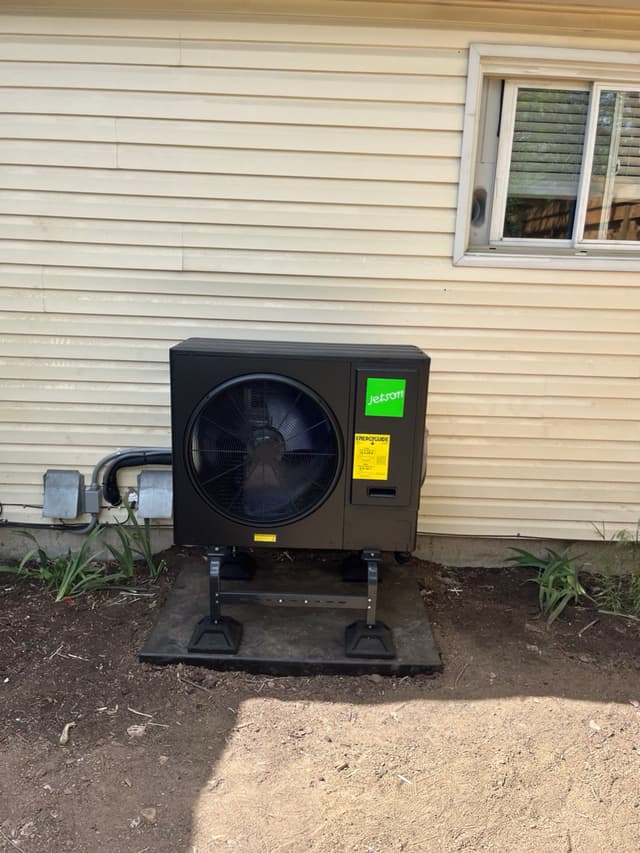 Black Jetson Air 3-ton heat pump condenser on an elevated pad stand against cream vinyl siding of a home in Thornton, CO, with electrical conduit and disconnect box mounted neatly on the wall beside it.