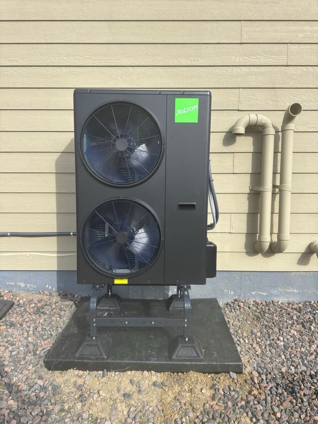 Jetson Air 4-ton dual-fan outdoor heat pump unit mounted on a black equipment stand over a rubber pad, set against gravel landscaping and beige lap siding of a home in Thornton, CO.