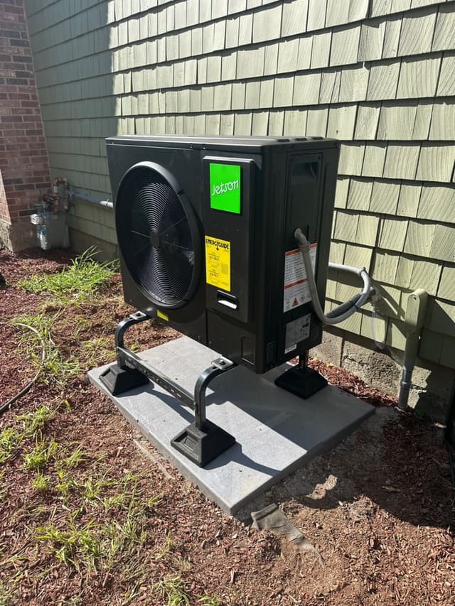Black Jetson Air 3-ton central ducted heat pump condenser installed against the gray shake siding of a house in Tyngsborough, MA, with refrigerant lines running to the exterior wall.