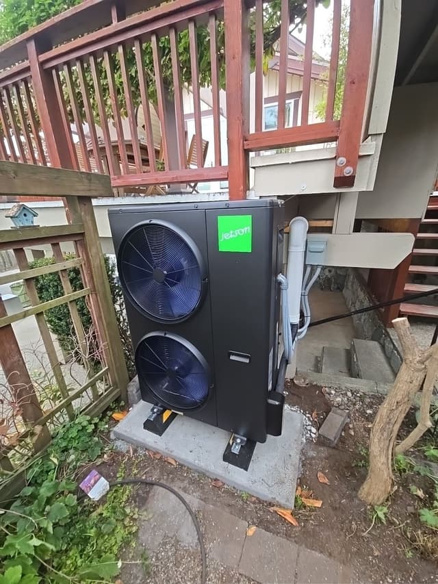 Black Jetson Air heat pump installed under a redwood deck beside a lattice fence in Hastings-Sunrise, Vancouver, with refrigerant lines running up to a disconnect box.
