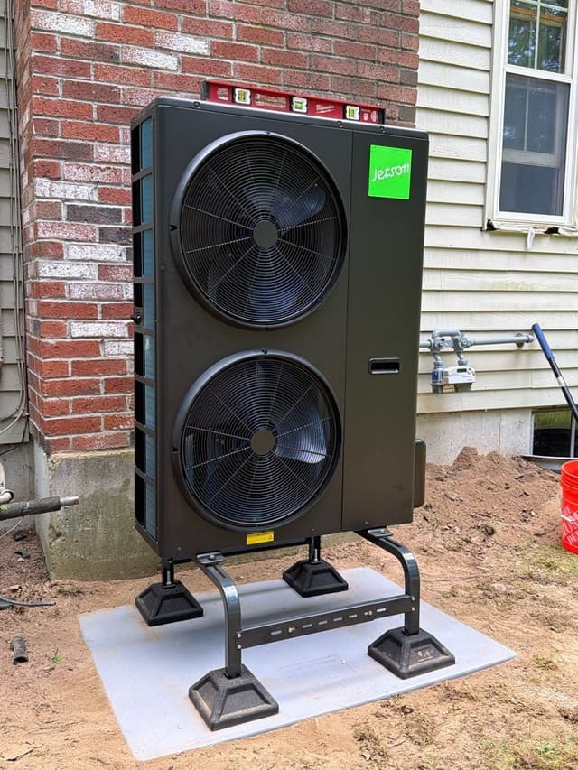 Jetson Air 5-ton central ducted heat pump outdoor unit installed in the corner between a brick wall and vinyl siding at a home in Walpole, MA.