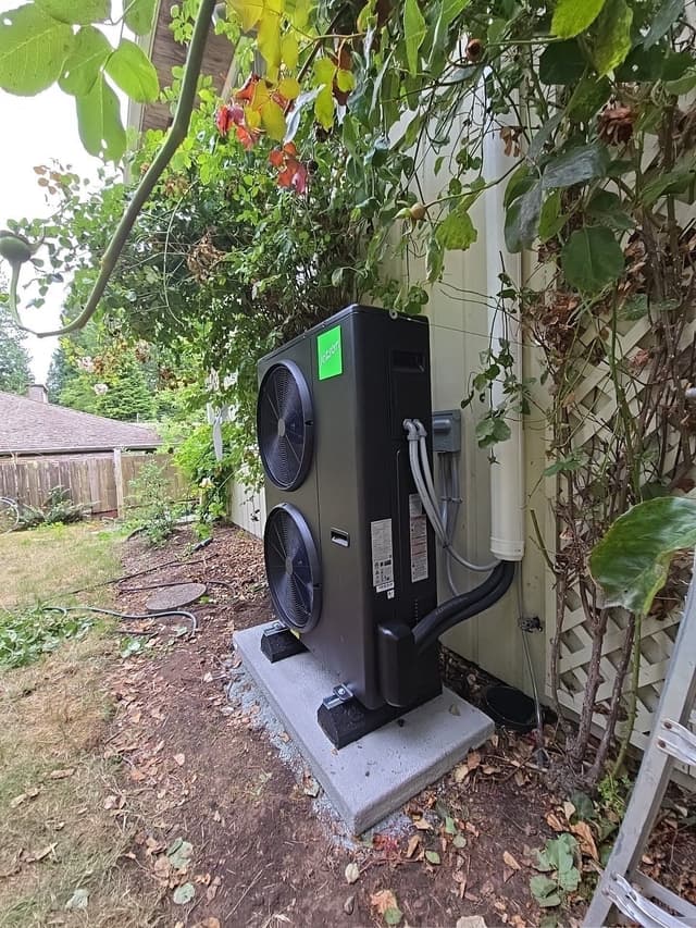 4-ton Jetson Air central ducted heat pump installed on a concrete pad against the side of a light-colored house in Eagle Harbour, West Vancouver, with refrigerant lines, an electrical disconnect box, a wooden fence, and overhanging tree branches visible.