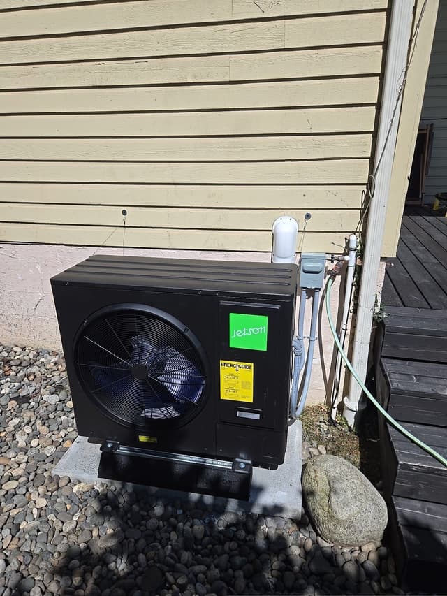 Black Jetson Air 3-ton heat pump installed on a concrete pad beside a dark wood deck on a home in West Bay, West Vancouver, with refrigerant lines running up cream-colored siding to a line set cover and disconnect box.