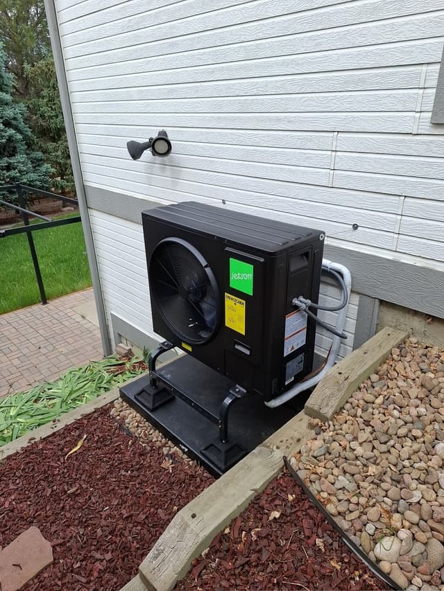 Jetson Air 3-ton heat pump condenser on a black riser pad along the side of a white vinyl-sided house, surrounded by river rock and red mulch with blue spruce trees in the background.